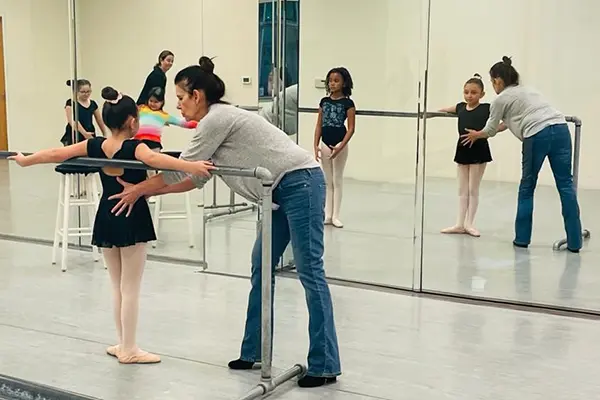 A woman assists young ballet students in front of a mirror.