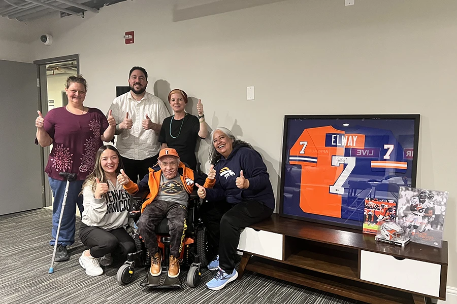 A group of people surround an older man in a wheelchair next to a table with Broncos football team mementos.