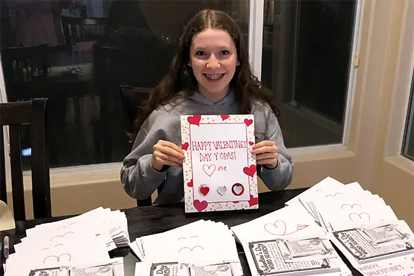 A teen girl holds a handmade Valentine's Day card with several others stacked on the table in front of her.