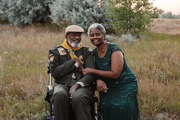 An older African American veteran in uniform sits in a field in a wheelchair, accompanied his wife