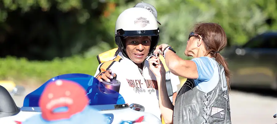A woman assists another woman with her motorcycle helmet.