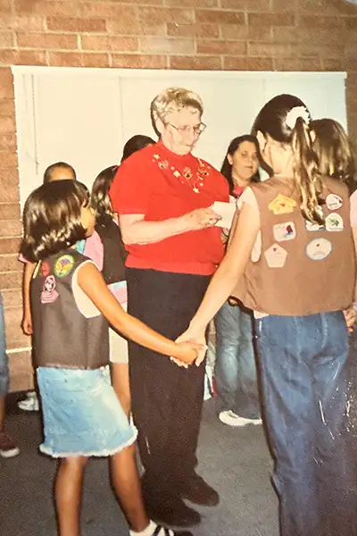 A woman stands in the center of a circle of girls wearing Girl Scout vests.