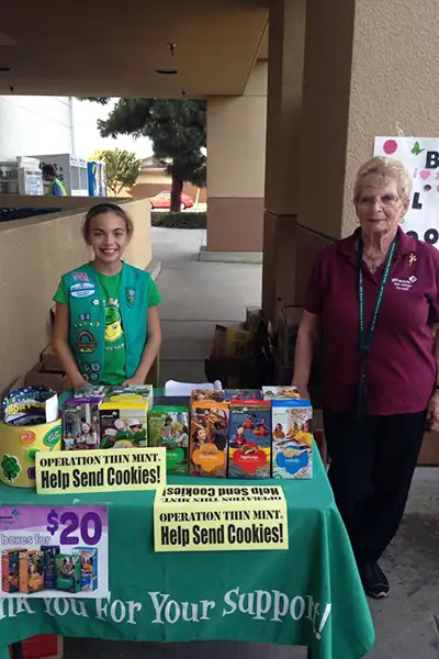 An older woman stands with a young Girl Scout at a cookie sales table.