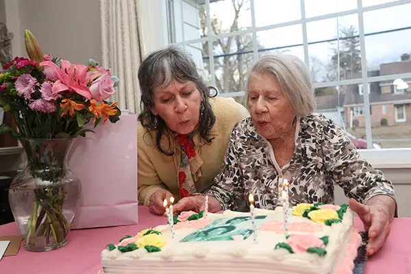 Two older women blow out candles on a birthday cake.
