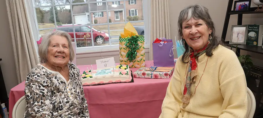 Two older women sit in front of a table with a birthday cake and presents.
