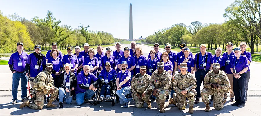 A large group of older people wearing purple shirts and a group of young men in combat fatigues in front of the Washington Monument