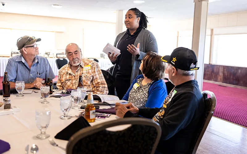 A woman talks with a group of men seated at a table.