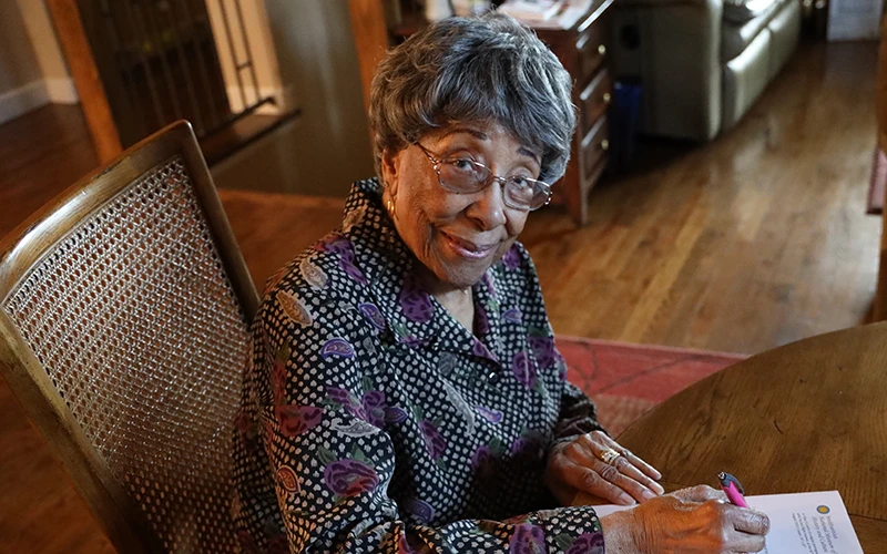 An older woman sits at a table while signing a paper