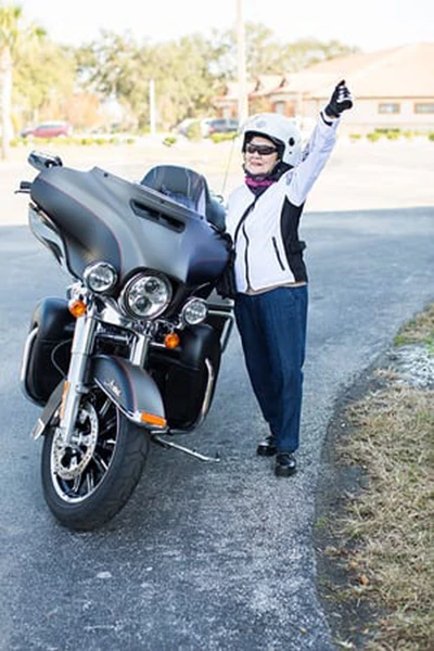 An older woman in full riding gear stands beside a large motorcycle