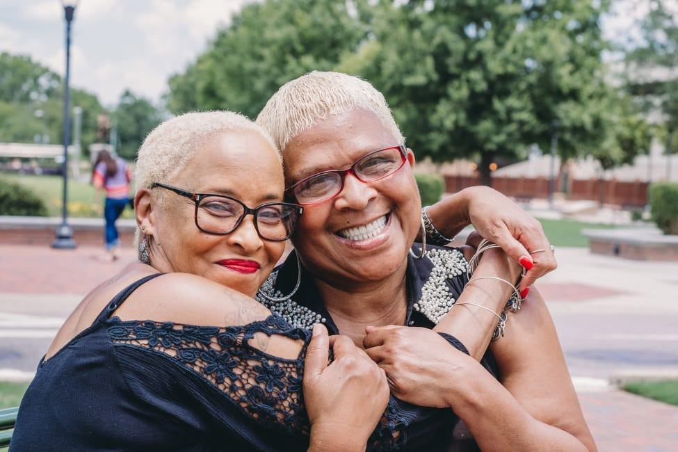Two older African American women smile at the camera while embracing each other