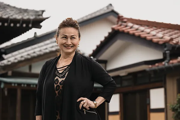 A Japanese woman smiles, standing in front of a house.
