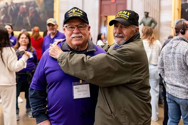 A older man in a Vietnam veteran hat hugs another veteran.