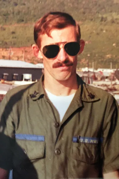 A man wearing sunglasses and a U.S. Navy working uniform from the 1960s.