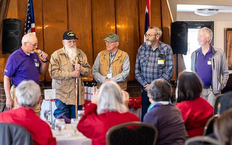Older men in veterans ball caps speak in front of an audience.