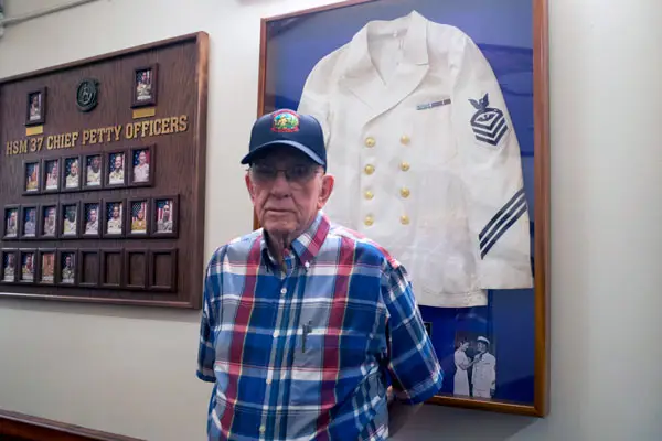 An older man wearing a WWII veteran ballcap stands in front of an exhibit of a Navy uniform