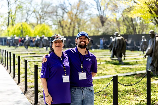 An older woman and younger man stand arm in arm at a memorial site.