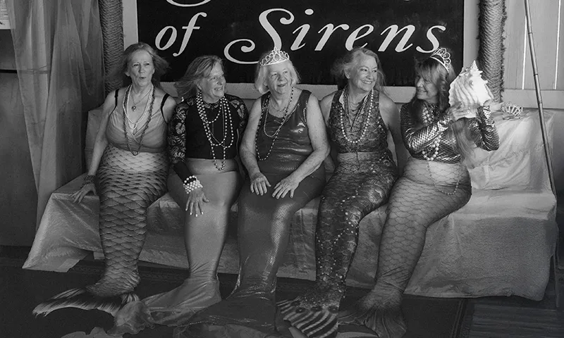 A group of older women wearing mermaid costumes sit on a bench