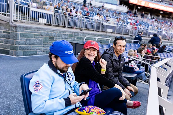 A woman and two men sit in a baseball stadium.