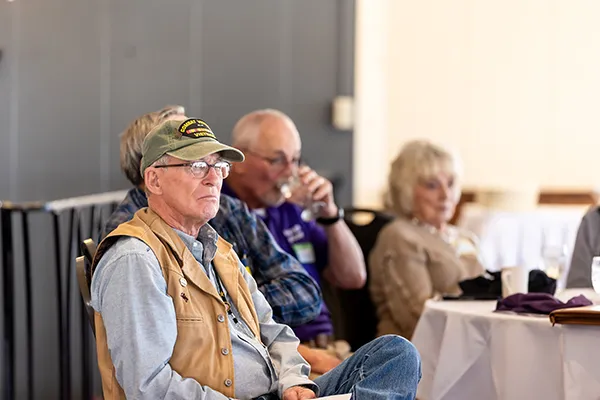 An older man sits at an event table with other people.