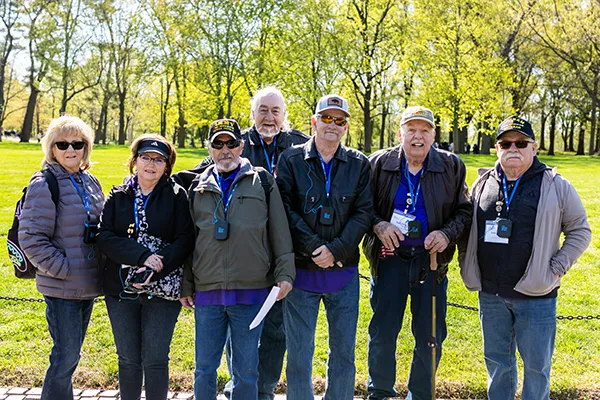 A group of older men and women stand outdoors by a tree-lined park.