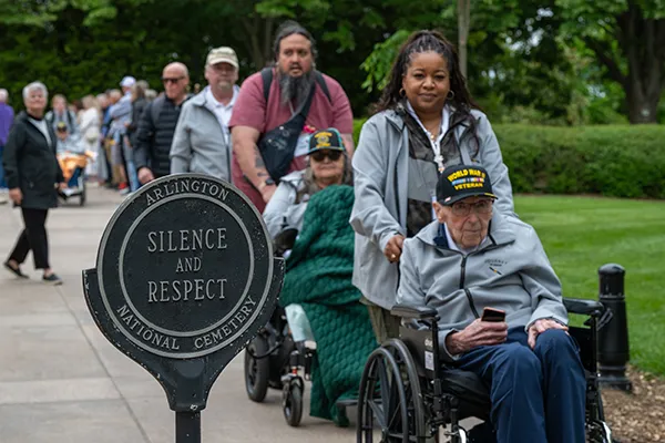 People in wheelchairs are escorted past a sign for Arlington National Cemetery