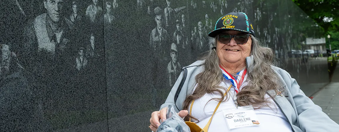 A woman in an Army veteran hat sits in front of the Mural Wall at the Korean War Veterans Memorial