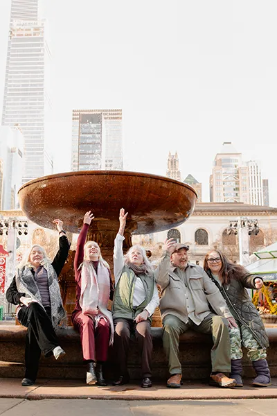 People sit together at the base of an outdoor fountain, raising their arms.