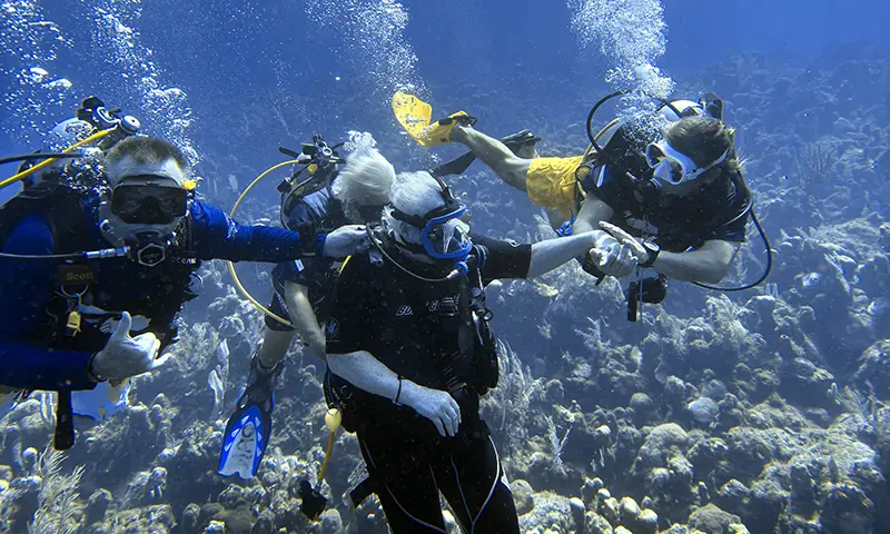 An older scuba diver is assisted by two other people
