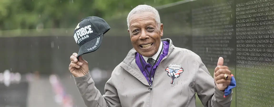 A man gives thumbs up while holding an<br />
Air Force ballcap in front of the Vietnam Veterans Memorial Wall.