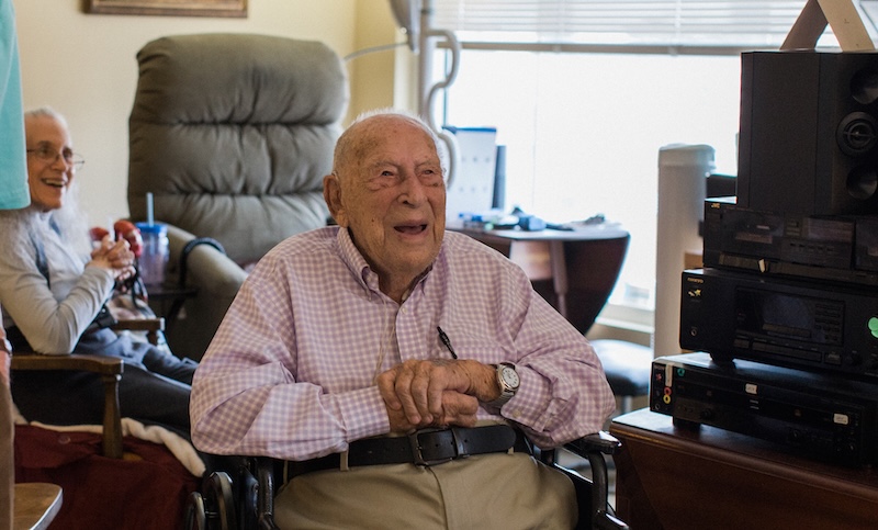 An older man in a wheelchair laughs while clasping his hands