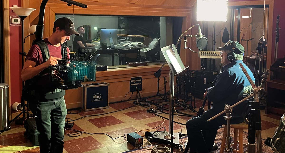 A man with a video camera records an older man singing a song in a recording studio