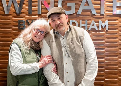 An older woman stands with her head on the shoulder of a younger man.