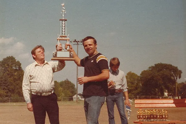 Two men hold up a baseball trophy in a 1980s photograph.