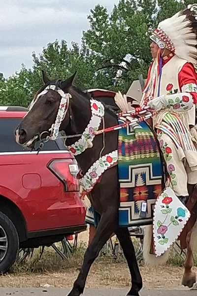 A Native person with wearing traditional regalia rides a horse past a truck.