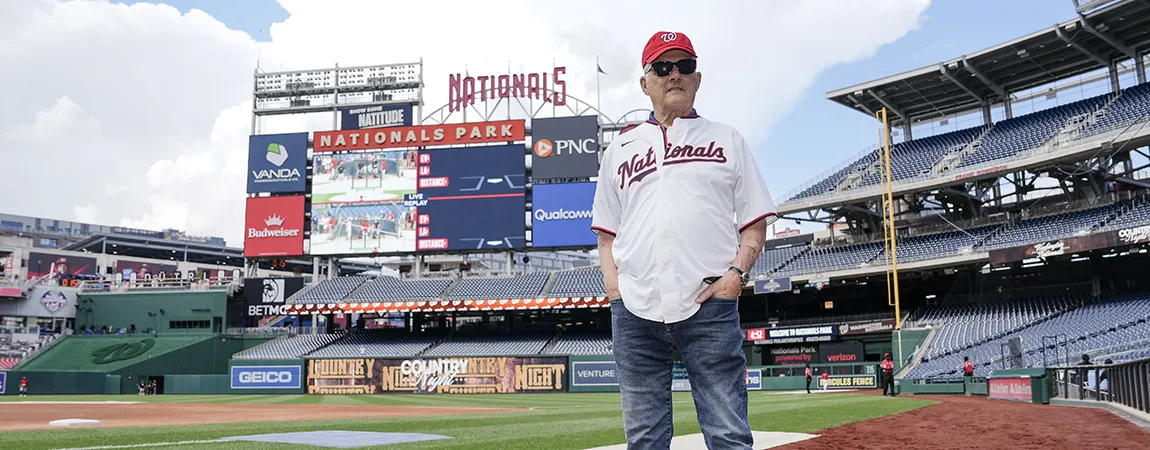 An older man wearing baseball gear stands on the Nationals baseball field.