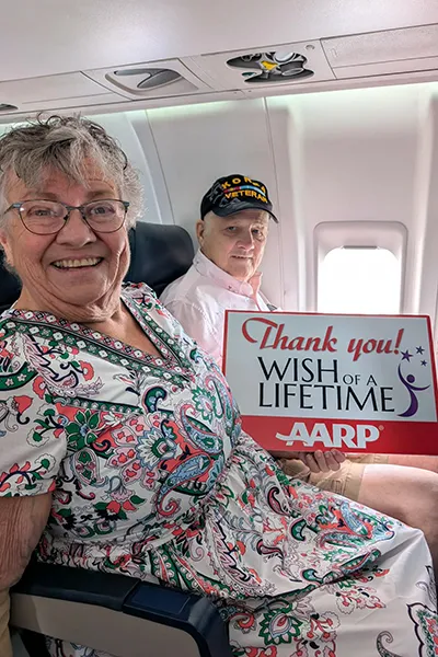A couple sits in airplane seats holding a Wish of a Lifetime from AARP sign.