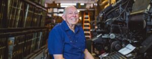 An older gentleman sits at an antique printing press and smiles at the camera.